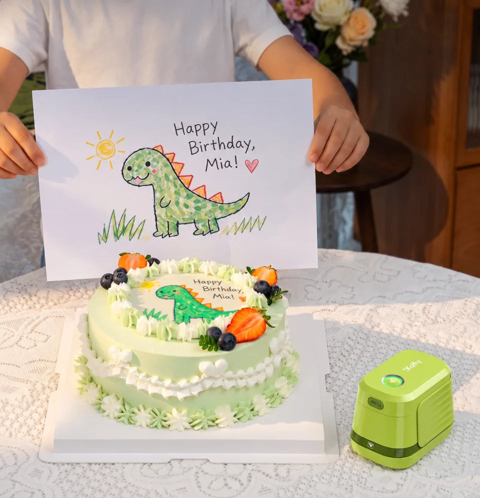 A dinosaur birthday cake beside the matching drawing and a green EdiBot printer.